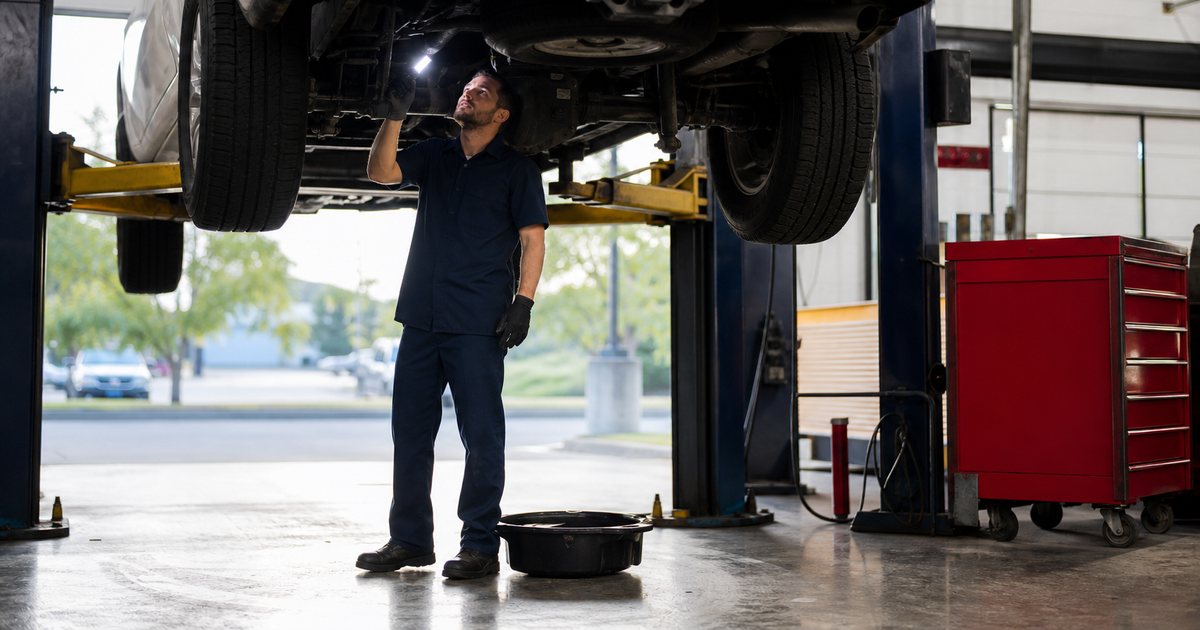 A mechanic in a navy work uniform standing beneath a white pickup truck raised on a two post lift in a clean repair bay, holding a portable work light up toward a suspension component, morning sunlight coming through an open roll up door, red tool cabinet and oil drain pan on the concrete floor